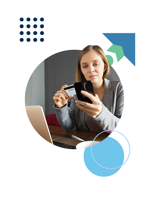 Woman in a grey blazer uses a smartphone to pay with a credit card at a desk with a laptop nearby, suggesting online shopping or payments.